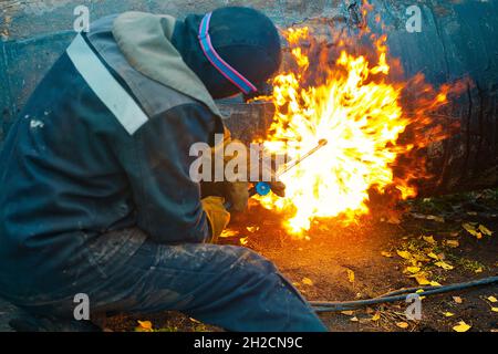 Der Schweißer sitzt in der Nähe des Rohres und schneidet es mit Hilfe des Cetylenschweißens. Ein Arbeiter auf der Straße schneidet Rohre mit großem Durchmesser und Funken und Feuer fliegen Stockfoto