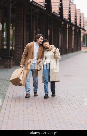 Liebevolles multiethnisches Paar in Mänteln, das die Hände hält, während es mit Einkäufen in der Nähe des Einkaufszentrums geht Stockfoto