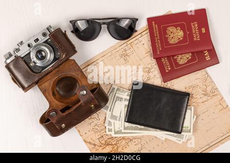 Weltkarte, zwei Pässe, Geld in einer schwarzen Ledertasche, eine alte Filmkamera in einem Lederetui und Sonnenbrille auf einem weißen Holztisch. Blick vom Abov Stockfoto