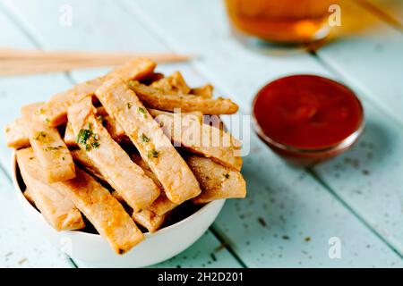 Nahaufnahme einer weißen Keramikschale mit frittierten Streifen Hühnchen auf einem blauen gedeckten Tisch neben einer Glasschüssel mit Ketchup Stockfoto