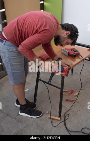 Junger Mann, der die Elektrowerkzeuge des Zimmermanns verwendet, um in seinem Haus Holzmöbel zu machen Stockfoto