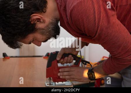 Junger Mann, der die Elektrowerkzeuge des Zimmermanns verwendet, um in seinem Haus Holzmöbel zu machen Stockfoto