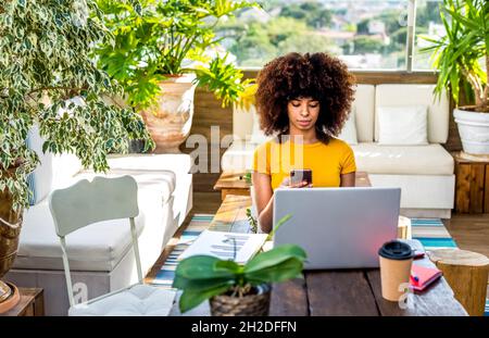 Junge, multiethnische Frau, die beim Start-up-Projekt arbeitet, sitzt auf einem Tisch im Coworking Space und hält ein Smartphone, um über ihre Arbeit zu diskutieren Stockfoto