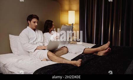 Mann mit Laptop in der Nähe einer jungen Freundin, die Zeitung las und sich im Hotelzimmer ausruhte Stockfoto