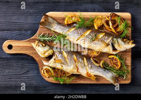 Gebratener Seebarsch-Fisch auf einem Holzbrett mit Meersalz, Dill, Zitrone auf einem schwarzen Holztisch, Draufsicht, Nahaufnahme Stockfoto