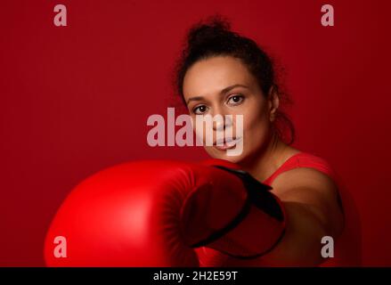 Nahaufnahme eines konzentrierten Sportfrauenboxers mit roten Boxhandschuhen, der direkt auf die Kamera trifft, isoliert über rotem Backgrou Stockfoto