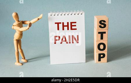 Der Text Stop Pain ist auf einem Notizbuch und Holzwürfeln geschrieben, neben einer Holzperson auf hellblauem Hintergrund. Medizinkonzept Stockfoto