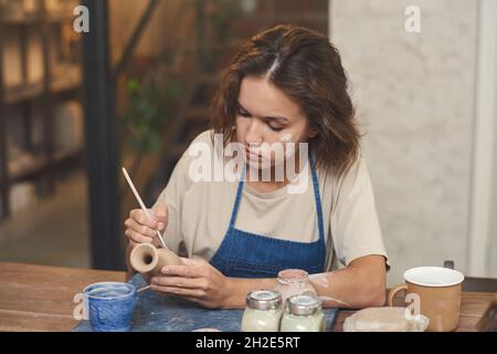 Konzentrierter junger kaukasischer Töpfer mit Schlamm auf der Wange, der am Schreibtisch sitzt und in der Werkstatt Tonvase malt Stockfoto