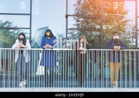 Business, Social Distancing, covid-19-Konzept. Junge schöne Gruppe von Menschen mit schwarzen Gesichtsmasken mit Handys mit Abstand in Glas Skyscrap Stockfoto
