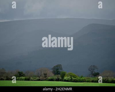 Brütende Hügel der Northern Pennines unter starken Wolken kontrastieren mit hellgrünem Vordergrundfeld und Bäumen am Flussufer in Cumbria, England, Großbritannien Stockfoto