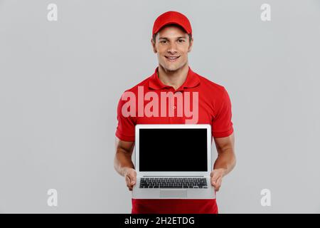 Der junge Mann, der in Uniform geliefert wurde, lächelte und zeigte einen Laptop, der über einer weißen Wand isoliert war Stockfoto