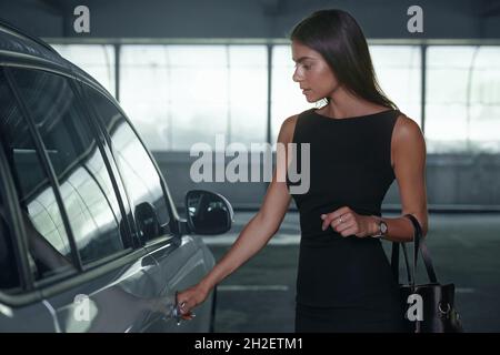 Schöne europäische Mädchen öffnen Tür ihres privaten Autos. Ernst junge Brünette Frau tragen schwarzes Kleid und Gold Handtasche. Konzept der erfolgreichen Mod Stockfoto