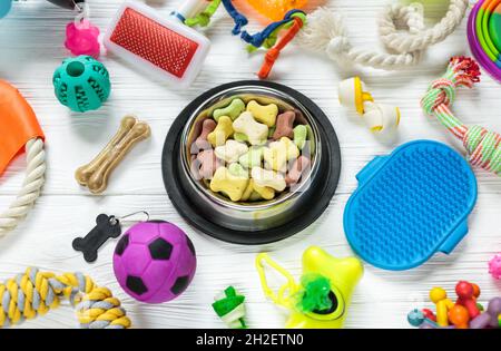 Verschiedene bunte Tierpflegezubehör: Schale, Knochen, Kugeln, Snacks auf weißem Holzhintergrund. Gummi- und Textilspielzeug für Hunde. Draufsicht, Stockfoto