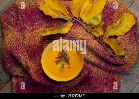 Herbstliches Food-Konzept, Kürbissuppe in einer Glasschale, verziert mit einem orangefarbenen Strickpullover und einem Zweig mit gelben Herbstblättern Stockfoto