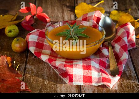 Kürbissuppe in einer Glasschüssel auf einem Holztisch, geschmückt mit bunten Herbstblättern und kleinen wilden Äpfeln Stockfoto