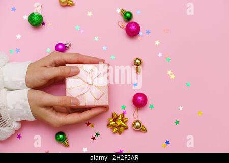 Frau Hände halten Geschenk, dekoriert mit Band auf rosa Hintergrund mit weihnachtskugeln und bunten Süßigkeiten Sterne, kopieren Raum. Flach liegend, Hände und Geschenkbox, Draufsicht. Weihnachtskonzept. Stockfoto