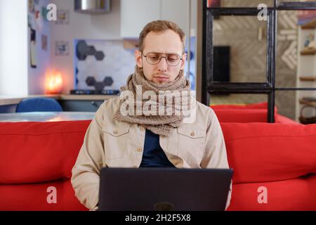 Ein Mann in einem Schal sitzt zu Hause mit einem Laptop auf der Couch. Erkältungen, Telearbeit, kaltes Wetter, Selbstisolierung Stockfoto