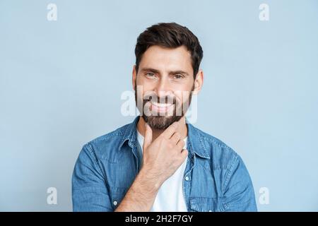 Porträt eines kaukasischen Mannes, der lächelt und den Bart auf dem Gesicht über der blauen Wand berührt Stockfoto