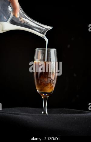 Vertikale Aufnahme eines Mannes, der Milch in ein Sektflötenglas voller Kaffee gießt; schwarzer Hintergrund Stockfoto
