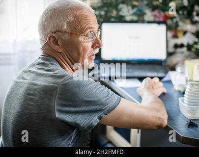 Älterer Mann sitzt am Tisch neben dem offenen Laptop. Alter Mann lernt am Computer zu arbeiten. Stockfoto