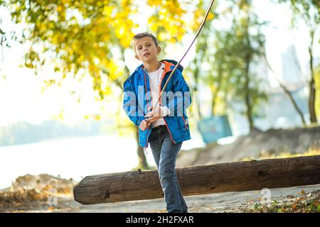 Ein Junge mit einem Ast läuft am Ufer des Flusses in der Stadt entlang. Ein Kind spielt in einem Stadtpark im Herbst vor dem Hintergrund der gelben fol Stockfoto