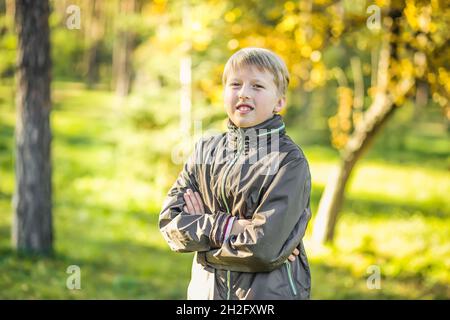 Hübscher Junge steht mit auf der Brust gefalteten Händen vor dem Hintergrund des Parks mit Bäumen mit gelben Blättern. Spaziergänge in der Stadt im Herbst. Stockfoto