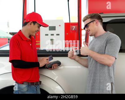 Tankstellenmitarbeiter in roter Uniform stehen lächelnd und wischen über das Zahlungsterminal mit einer Mockup-Kreditkarte. Der kaukasische Fahrer mit Sonnenbrille hebt beide Daumen nach oben Stockfoto