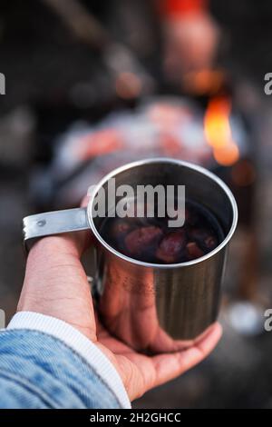 Frau Hand halten Rose Hip Tee in Metall-Becher gegen Lagerfeuer. Umweltfreundlicher Tourismus, nachhaltiger Lebensstil. Stockfoto