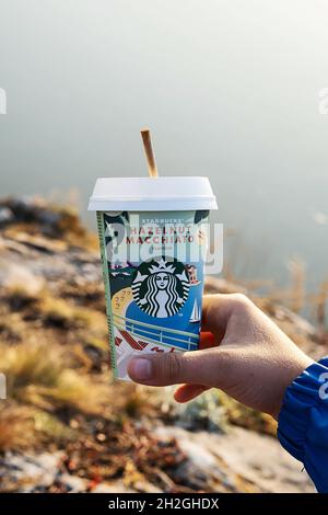 Kemerowo, Russland - 17. Oktober 2021: Mann hält Starbucks-Kaffee auf landschaftlichem Hintergrund. Haselnuss Macchiato to go Kaffeetasse, Nahaufnahme. Stockfoto