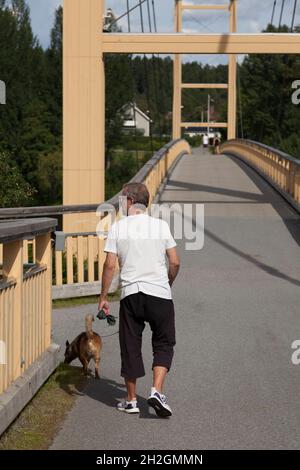 Skelleftea, Norrland Schweden - 18. August 2020: Älterer Mann, der seinen Hund über eine Holzbrücke führt Stockfoto