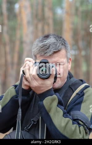 Porträt eines Fotografen. Ein Mann mittleren Alters schaut durch die Linse seiner Kamera in die Kamera. Verschwommene Pinien im Hintergrund. Stockfoto