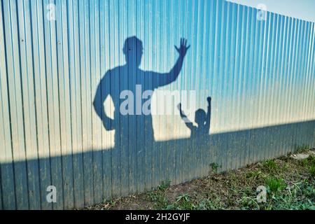 Schatten von positivem Vater und kleinem Kind mit erhobenen Händen auf Metallzaunrahmen im Sommerhausgarten bei Sonnenuntergang Stockfoto