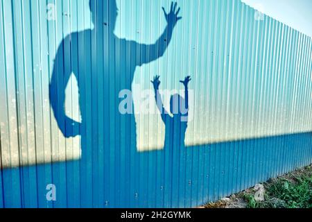 Schatten von positivem Vater und kleinem Kind mit erhobenen Händen auf Metallzaunrahmen im Sommerhausgarten bei Sonnenuntergang Stockfoto