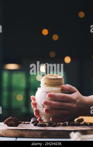 Hausgemachter Dalgona-Kaffee in einem Einmachglas. Hände eines Mädchens, das den Dalgona Kaffee hält. Stockfoto