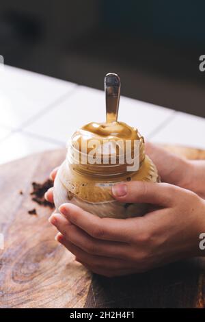 Hausgemachter Dalgona-Kaffee in einem Einmachglas. Hände eines Mädchens, das den Dalgona Kaffee hält. Stockfoto