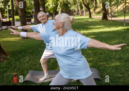 Lächelnder älterer Mann sieht die Partnerin an, die gemeinsam im Stadtpark Yoga-Übungen macht Stockfoto