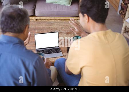 Sohn und älterer Vater mit birazialen Erwachsenen halten Tassen Kaffee und führen einen Videoanruf auf dem Laptop durch Stockfoto