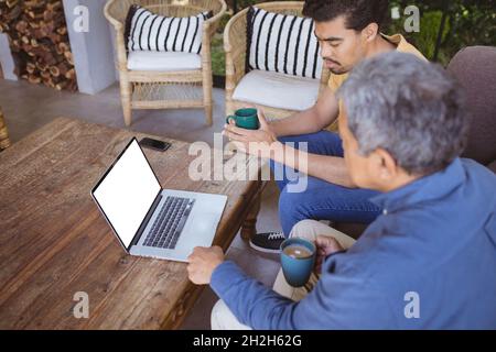 Sohn und älterer Vater mit birazialen Erwachsenen halten Tassen Kaffee und führen einen Videoanruf mit dem Laptop durch Stockfoto