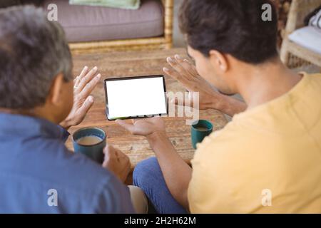 Sohn und älterer Vater mit birazialen Erwachsenen halten Tassen Kaffee und führen einen Tablet-Videoanruf durch Stockfoto