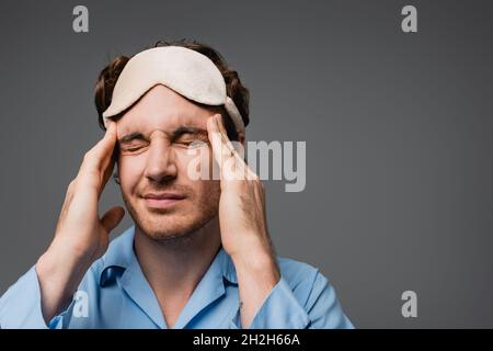 Junger Mann in Pyjama und Schlafmaske mit Kopfschmerzen isoliert auf grau Stockfoto