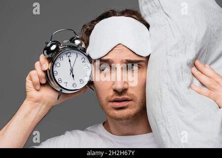 Junger Mann in Schlafmaske mit Kissen und Wecker isoliert auf grau Stockfoto
