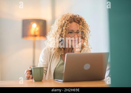 Schöne kaukasische junge Frau mit lockigen Haaren und Brillen, die Kaffee trinkt und auf den Laptop-Bildschirm schaut. Frau, die Kaffee trinkt, während sie einen Laptop benutzt Stockfoto