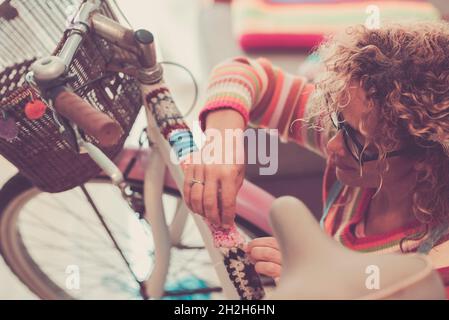 Frau, die Fahrradstange mit Wollstoff schmückt. Frau mit lockigen Haaren und Brillen dekorieren oder modifizieren ihr Fahrrad mit gestricktem Wollstoff o Stockfoto