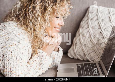 Glückliche junge Frau, die vor dem Laptop auf dem Sofa liegt und die Hände zu Hause im Wohnzimmer zusammengeschleppt hat. Geschäftsfrau in der Freizeit, die von zu Hause aus arbeitet. Kaukasischer wo Stockfoto