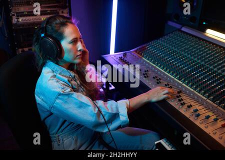 Junge Frau mit Kopfhörern, die Musik hört und auf der Musiktastatur tippt, um ein Lied aufzunehmen Stockfoto