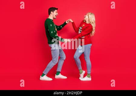 Foto von verträumten Paar in der Liebe Tanz halten Hände tragen hässlichen Pullover Jeans Sneakers isoliert roten Hintergrund Stockfoto