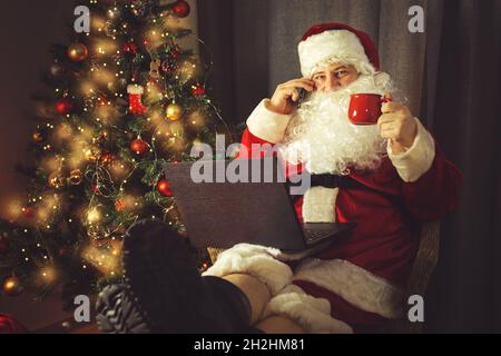 Der authentische Weihnachtsmann arbeitet an einem Laptop und telefoniert. Inneneinrichtung. In Erwartung von Weihnachten und Neujahr. Stockfoto