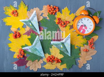 Papierflugzeuge aus Euro-Banknoten und Wecker auf blauem Hintergrund mit roten und gelben Blättern und orangefarbenen Beeren. Herbstzeit für Reisen. T Stockfoto