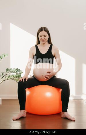 Eine Schwangere krümmt sich vor Schmerzen, ihr Arm liegt auf ihrem Bauch auf einem orangefarbenen Übungsball bei ihr zu Hause am Abend. Komplizierte Schwangerschaft. Stockfoto