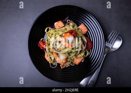 Nudeln mit Meeresfrüchten, getrockneten Tomaten, Kapern und roten Zwiebeln. Hausgemachte Pasta mit Garnelen, Lachs (Forelle) und Pesto-Sauce. Schwarzer Hintergrund, schwarzer p Stockfoto